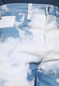 Jeans i denim med ett mönster av molnig himmel i nyanser av ljusblått och vitt. Inkluderar en framficka och metallknappdetaljer.