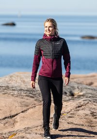 Maroon and black zip-up jacket with padded shoulders, black accents, and a hood; paired with black pants and black footwear; outdoor setting.