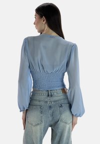 Hellblaue, durchsichtige Bluse mit voluminösen Ärmeln und einem gerafften Bund. Über abgewetzten Denim-Jeans getragen, mit detaillierten Akzenten an den hinteren Taschen.