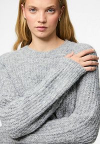 Blonda sieviete ar ziliem acīm, tērpusies mīkstā, gaiši pelēkā adītajā džemperī, ar kreiso roku tur pašu labo plecu.