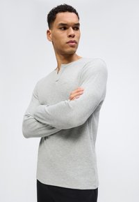 Άνδρας με σταυρωμένα χέρια φοράει ανοιχτό γκρι μακρυμάνικη μπλούζα henley, κοιτάζοντας προς το πλάι με φόντο απλό λευκό.