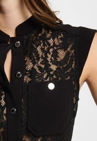 Zwarte blouse met kant overlay met bloemenpatronen, een effen zwarte kraag en knopen. Inclusief een voorzak met een zilveren accent.