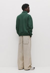 Grøn sweatshirt med høj krave, båret sammen med oversized beige bukser, der har to store lommer på bagsiden. Sorte og sølvfarvede sko fuldender looket.