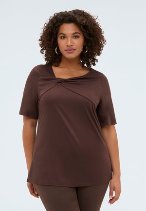 Kvinde med krøllet hår iført guldfarvede hoops-øreringe og en brun top med korte ærmer og en snoet halsudskæringsdetalje.