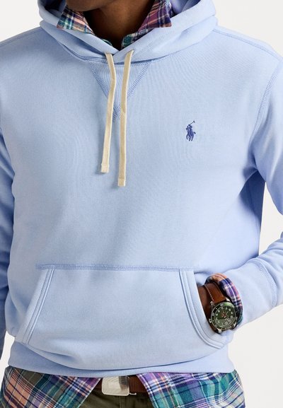 Helle blaue Kapuzenjacke mit einer Fronttasche, Zugkordel am Ausschnitt und lila Logo. Darunter trägt er ein grünes Karohemd. Eine braune Armbanduhr ist sichtbar.