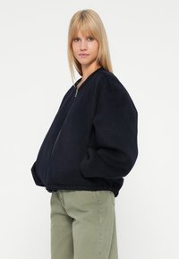 Joven con cabello rubio liso, vistiendo una chaqueta negra de gran tamaño con cremallera y pantalones verde claro, con las manos en los bolsillos de la chaqueta, fondo neutro.