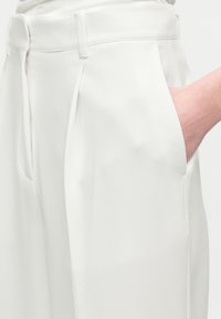 Main droite insérée dans la poche latérale d’un pantalon blanc taille haute, cintré, avec passants de ceinture et plis devant.