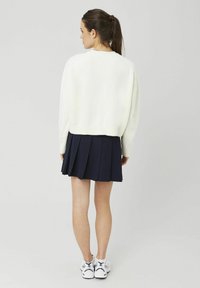 Femme avec une queue de cheval portant un pull blanc, une jupe plissée marine et des baskets blanches, debout de dos sur un fond uni.