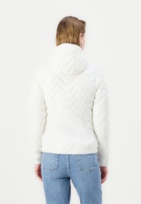 Giacca bianca trapuntata con un motivo a onde testurizzato, cappuccio e polsini aderenti, abbinata a jeans di denim blu.