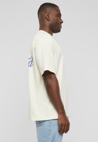 Beige short-sleeve t-shirt with a relaxed fit and a blue graphic on the back. Made of soft fabric, featuring a round neckline and straight hem.