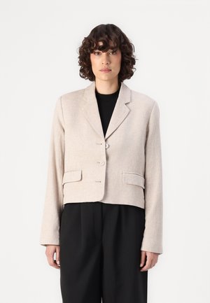 Gemiddeld beige blazer met een textuurafwerking, twee voorzakken, gelapte revers en drie knopen. Draag het over een zwarte top en broek.
