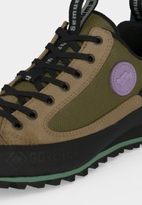 Υπαίθριο πεζοπορικό παπούτσι σε ελιά πράσινο και ταμπ με μαύρα κορδόνια, μωβ στρογγυλό λογότυπο, και μαύρη σόλα με πράσινη ρίγα και ετικέτα Gore-Tex.