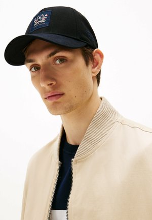 Schwarze Baseballmütze mit aufgesticktem Logo, kombiniert mit einer beigen Bomberjacke mit ribbeltem Kragen und metallischem Reißverschluss, getragen über einem marineblauen Shirt.