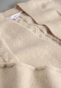 Zachte beige gebreide cardigan met geribde kraag en kabelbreisel details aan de voorkant, plat gelegd op een lichte ondergrond.