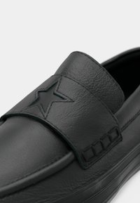 Svart lær sko med en fremre stropp som har preget stjernesmykke, glatt tekstur og sydd detaljer langs siden.