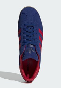 Blå ruskinds Adidas sneakers med røde striber og indvendig, snøret op, set ovenfra på en lys baggrund.