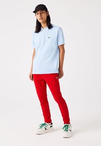 Lyseblå polo t-shirt og røde sweatpants. Poloen har et grønt logo; sweatpants har logo på benet. Hvide sportssko med grønne snørebånd.