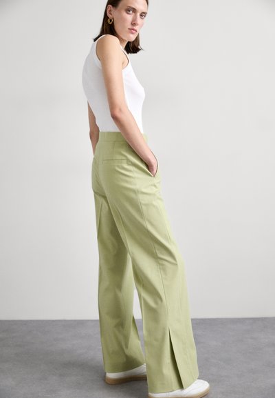 Lichtgroene wijde broek met twee zijzakken, een achterzak en een kleine split aan de zoom. Gekleed met een witte mouwloze top en sneakers.