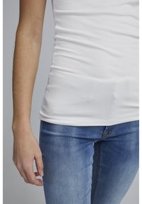 Personne portant un t-shirt blanc uni ajusté et un jean bleu, avec le bras gauche détendu le long du corps sur un fond neutre.
