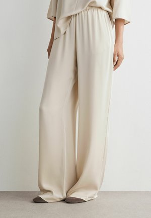 Beige bukser med vide ben og elastisk talje, båret med matchende top, der dækker grå spidse sko på en neutral baggrund.