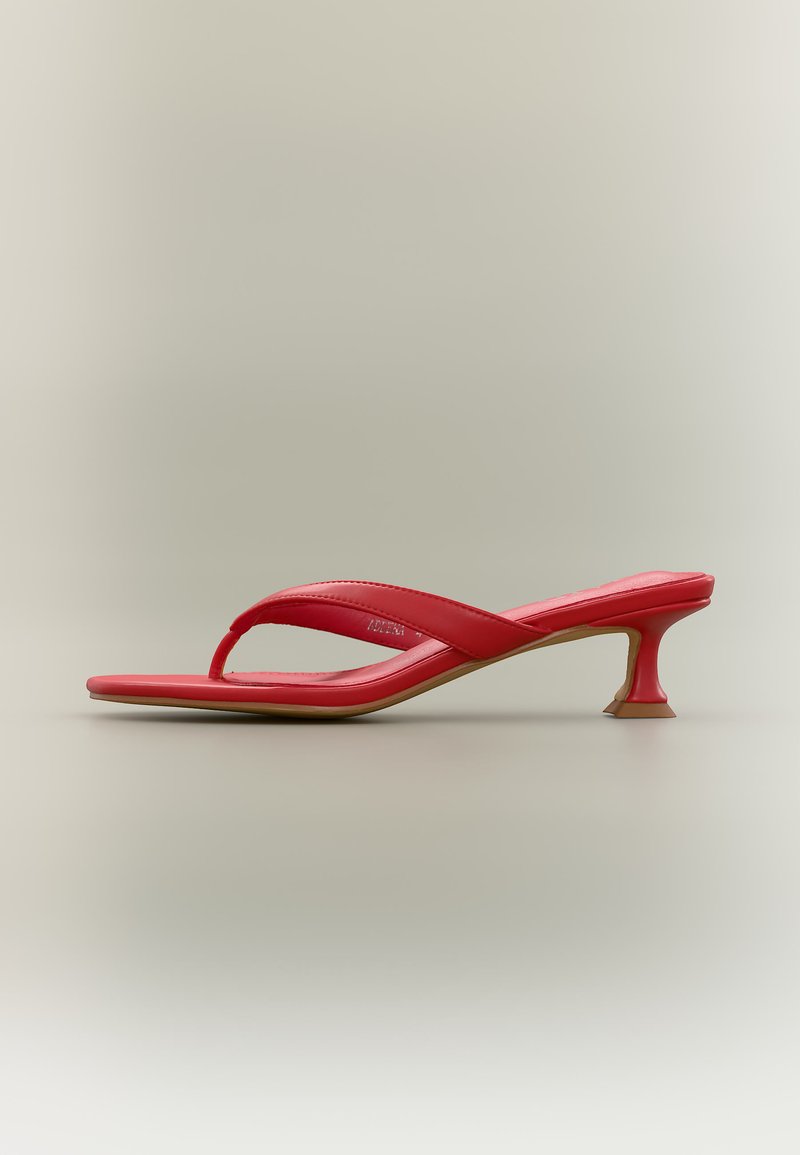 Sandale à talon rouge avec lanière en forme de tanga, bout carré, petit talon incurvé et brides rembourrées sur un fond clair uni.