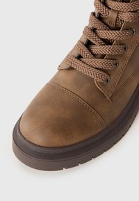 Bottine en cuir brun avec des lacets épais tressés, bout rond, couture détaillée et semelle en caoutchouc noir striée sur fond blanc.
