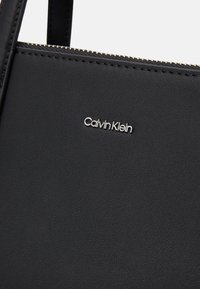 Borsa nera in ecopelle con chiusura a zip, caratterizzata da un logo "Calvin Klein" in argento e dettagli di cucitura fine. Texture liscia.