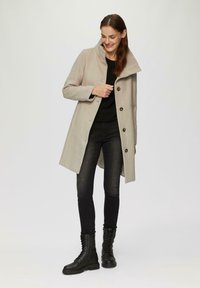 Beige wollen blend jas met een hoge kraag, vijf knopen en een rechte snit. Gedragen over een zwarte trui en gecombineerd met zwarte skinny jeans en veterschoenen.