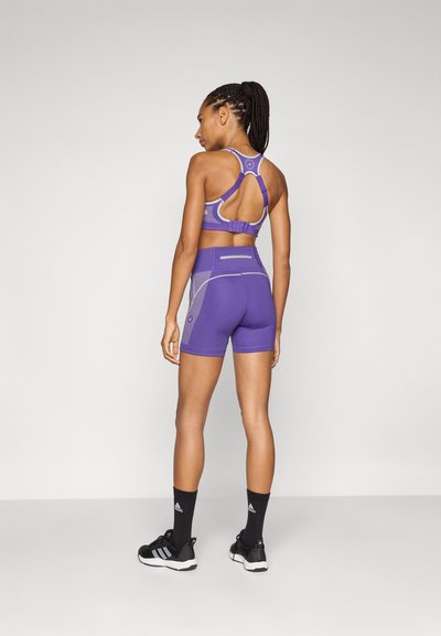 Lila sportmelltartó és magas derekú rövidnadrág geometriai mintákkal. Kivágásos dizájnnal és kontrasztos varrással. Fekete sportcipővel viselve.