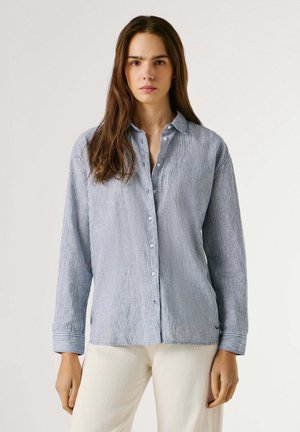 Junge Frau mit langen braunen Haaren trägt ein blau-weiß gestreiftes Hemd mit Knöpfen und cremefarbene Hose, steht vor einem neutralen Hintergrund.