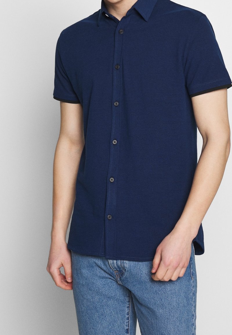 Polo a maniche corte color blu navy realizzato in tessuto morbido, con chiusura frontale a bottoni, colletto discreto e rifiniture a contrasto sulle maniche.