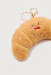 Porte-clés en forme de croissant en peluche avec un visage souriant brodé et un anneau en métal doré sur fond blanc.