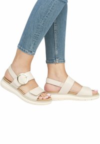 Sandales beiges à lanières avec un accent métallique circulaire, semelle rembourrée et lanières texturées, posées sur un jean en denim.