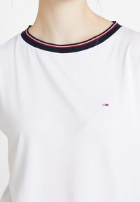 Bílý tričko s kulatým námořnickým límcem, zdobené červenými a bílými pruhy; malé logo umístěné na levé straně poblíž výstřihu.