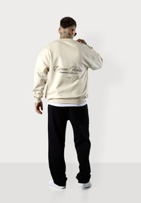 Sweatshirt crème avec texte au dos, associé à un pantalon noir et des baskets blanches. Le matériau semble doux et décontracté, avec une coupe ample.