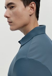 Polo in un morbido colore blu tenue con collo. Il tessuto sembra liscio e aderente, mostrando un design contemporaneo.