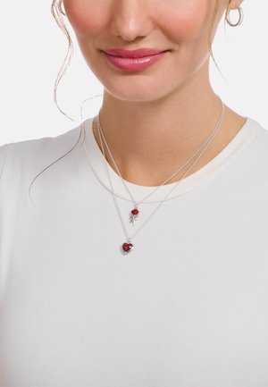 Silberne geschichtete Halsketten mit einem roten Herz-Anhänger und einem Rosen-Anhänger. Die Trägerin hat helle Haut und trägt ein schlichtes weißes Oberteil.