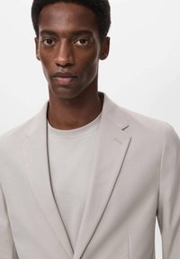 Beige tailored blazer with a subtle sheen, notched lapels, and a light gray crew neck t-shirt underneath. Smooth fabric and clean lines.