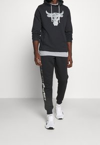 Uomo che indossa un felpa nera con logo di toro e pantaloni della tuta neri con scritto "Sweat Respect", abbinati a sneakers bianche Nike.