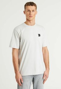 Camiseta de color gris claro, hecha de algodón, con cuello redondo, mangas cortas y un pequeño parche negro con el logotipo en el lado izquierdo del pecho.