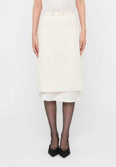 Jupe crayon crème à design superposé, texture lisse, ceinture avec détails métalliques. Portée avec des collants noirs transparents et des escarpins à bout pointu.