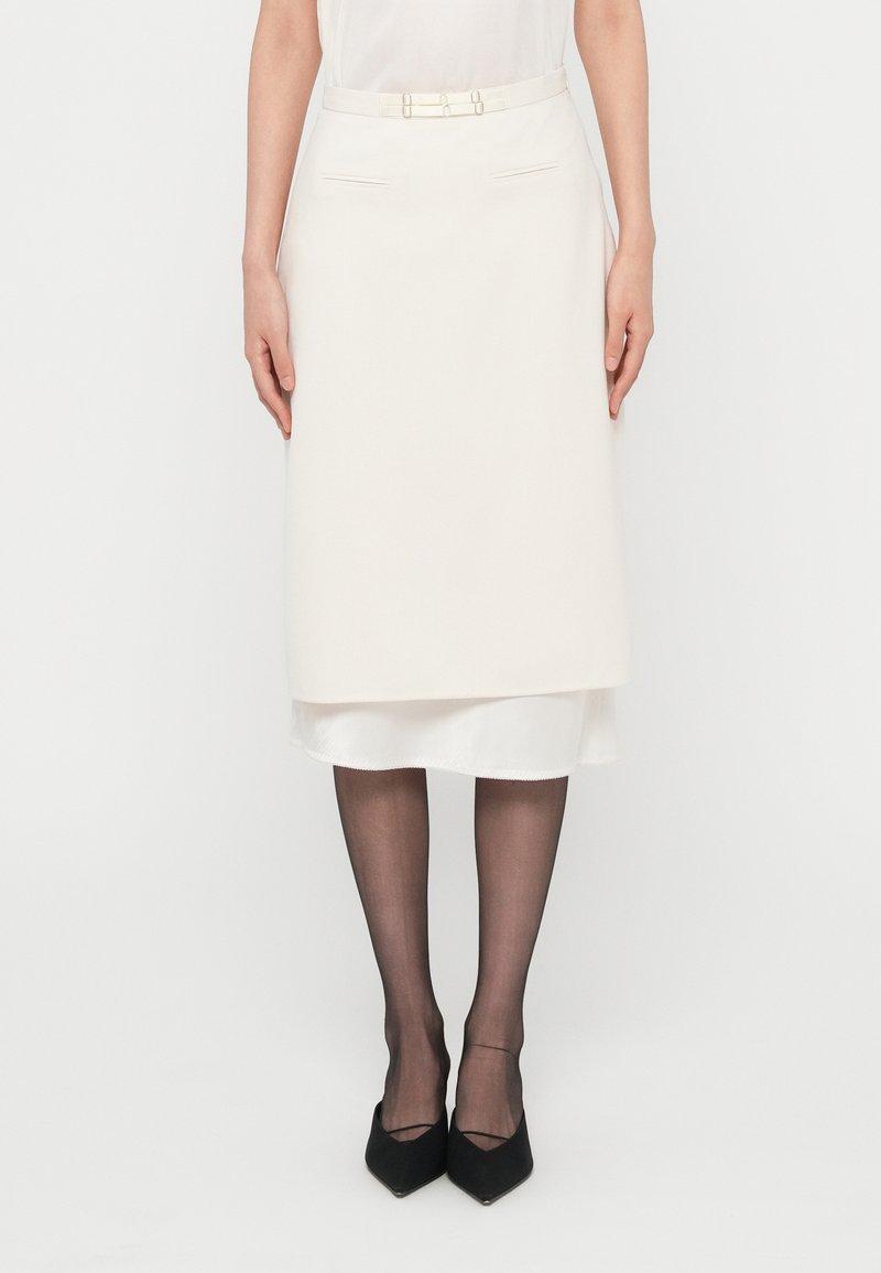 Jupe crayon crème à design superposé, texture lisse, ceinture avec détails métalliques. Portée avec des collants noirs transparents et des escarpins �à bout pointu.