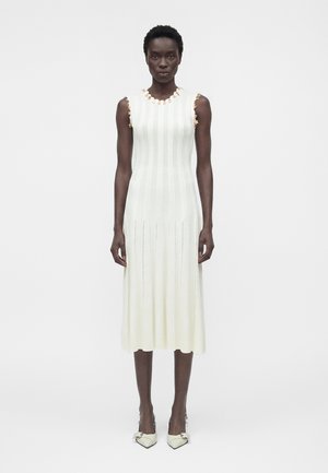 Femme portant une robe midi blanche sans manches en maille côtelée avec un galon floral au niveau du décolleté et des emmanchures, associée à des escarpins blancs pointus.