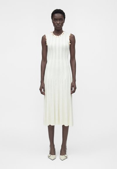 Femme portant une robe midi blanche sans manches en maille côtelée avec un galon floral au niveau du décolleté et des emmanchures, associée à des escarpins blancs pointus.