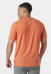 Krátký rukáv oranžového trička z hladkého materiálu, s kulatým výstřihem a jemnými detaily šití. Nosí se s šedými šortkami.