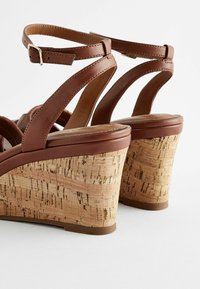 Sandali con zeppa in pelle marrone con tacco in sughero, dotati di cinturini regolabili alla caviglia e superficie liscia. Design semplice con dettagli di cuciture sottili.