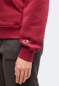 Gros plan sur la main d'une personne qui porte un sweat-shirt rouge avec des poignets côtelés et un petit logo Champion sur la manche, associé à un pantalon foncé.