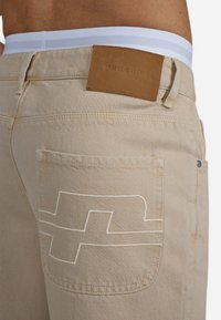 Jeans bej cu un buzunar mare la spate, cu cusături albe și o etichetă din piele inscripționată "PEGADOR". Talie cu dungi albe și gri.
