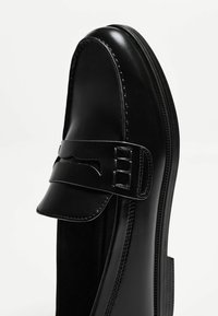 Black leather loafer featuring a rounded toe, a decorative strap with cutout detailing, and subtle stitching along the edges.