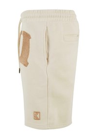 Beige sweat shorts made of soft fabric, featuring a side pocket, an embroidered brown letter "D," and an embossed logo patch.